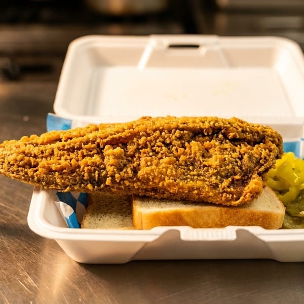 Crispy fried fish, white bread, and pickles in a white takeout container on a metal counter.