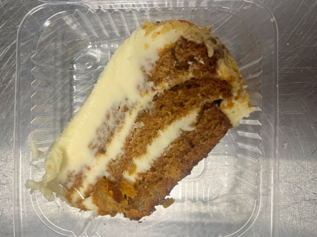 A slice of carrot cake with cream cheese frosting in a clear plastic container.