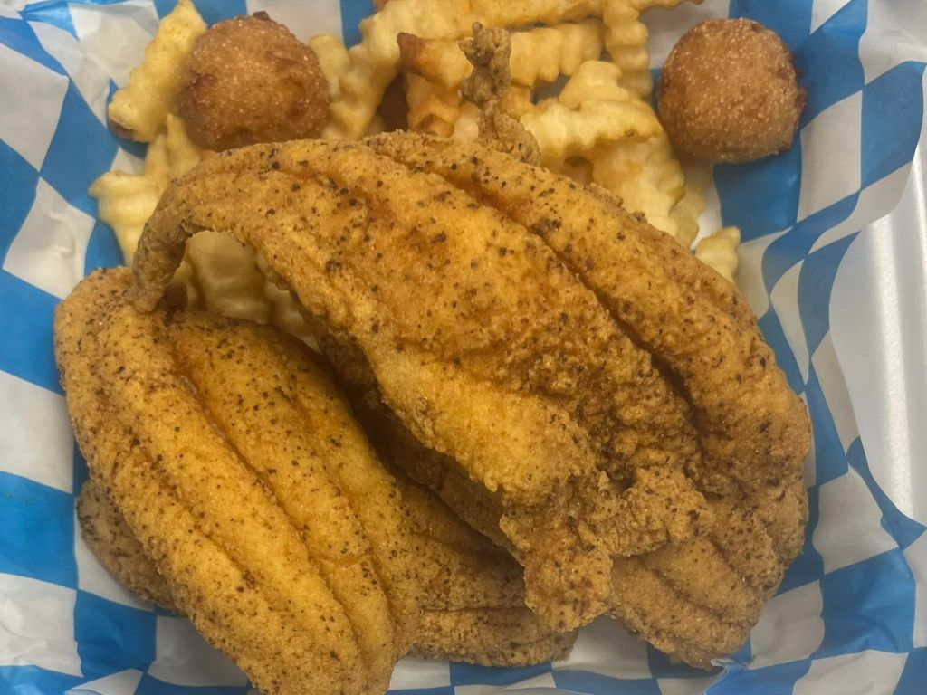 A serving of crispy fried catfish fillets accompanied by crinkle-cut fries and two hushpuppies, arranged on a blue and white checkered tray.